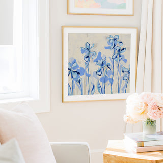 a cozy living room with a white sofa, a framed artwork of blue flowers on the wall, and a vase of pink flowers on a wooden table.