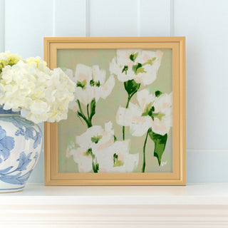 a framed painting of white flowers with green stems, placed on a white shelf next to a blue and white vase with white flowers.