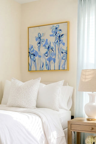 a serene bedroom scene with a bed adorned with white linens and a framed painting of blue flowers hanging above it. The painting adds a touch of color and life to the otherwise neutral-toned room.
