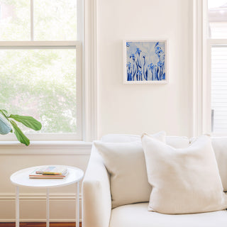 a cozy living room with a white sofa, a small round table, and a framed artwork on the wall. The room is bright and airy, with natural light coming in through a window.