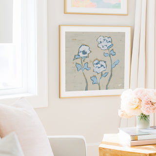 a cozy living room with a framed painting of blue flowers hanging on the wall, a vase of pink flowers on a wooden table, and a stack of books on the table.