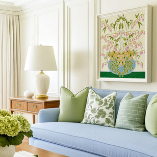 a cozy living room with a blue sofa adorned with green and white throw pillows, a white lamp on a wooden side table, and a framed floral artwork hanging on the wall above the sofa.