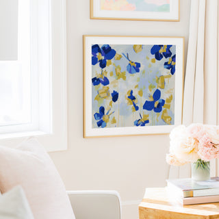 a cozy living room with a white sofa, a framed artwork of blue and yellow flowers on the wall, and a vase of pink flowers on a wooden table.