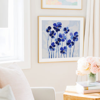 a cozy living room with a white sofa, a framed painting of blue flowers on the wall, and a vase of pink flowers on a wooden table.