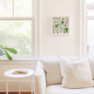 a cozy living room with a white sofa, a small white side table, and a framed picture of white flowers hanging on the wall.