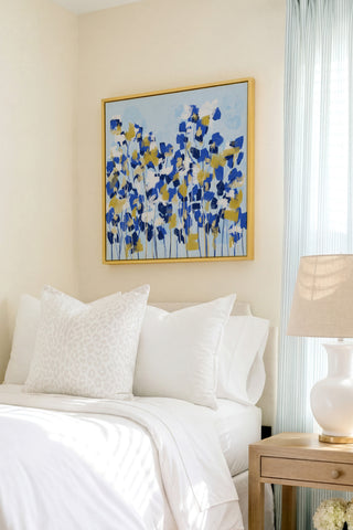 a serene bedroom scene with a bed adorned with white pillows and a blue and white floral painting hanging above it. The painting adds a touch of color and life to the otherwise neutral-toned room.