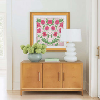 a wooden cabinet with a vase of green flowers on top, and a white lamp on the right side. The cabinet is situated in a room with a white wall and a framed picture hanging above it.