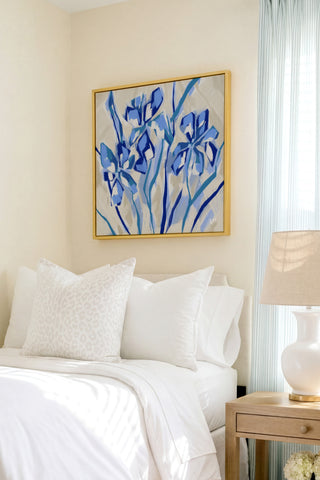 a serene bedroom scene with a bed adorned with white linens and a framed painting of blue flowers hanging above it. The painting adds a touch of color and life to the otherwise neutral-toned room.