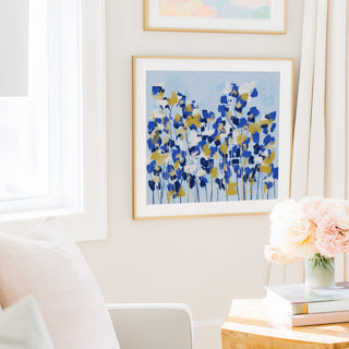 a cozy living room with a white sofa, a framed painting of blue and yellow flowers hanging on the wall, and a vase of pink flowers on a wooden table.