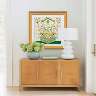 a wooden cabinet with a vase of green flowers on top, and a white lamp on the right side. The cabinet is situated in a room with white walls and a wooden floor.