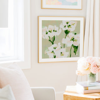 a cozy living room with a large framed painting of white flowers hanging on the wall, a white chair, a small table with a vase of pink flowers, and a stack of books.