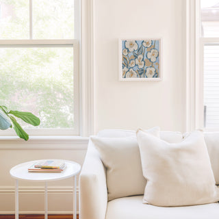 a cozy living room with a white sofa, a small round table, and a framed artwork on the wall. The room is bright and airy, with natural light coming in through a window.