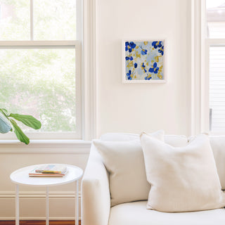 a cozy living room with a white sofa, a small round table, and a framed artwork on the wall. The room is bright and airy, with natural light coming in through a window.