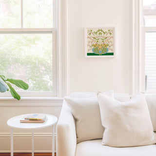 a cozy living room with a white sofa, a small round table, and a framed artwork hanging on the wall. The room is bright and airy, with natural light coming in through a window.