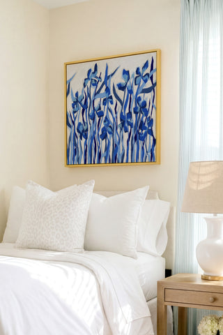 a serene bedroom scene with a bed adorned with white linens and a framed painting of blue flowers hanging above it. The painting adds a touch of color and life to the otherwise neutral-toned room.