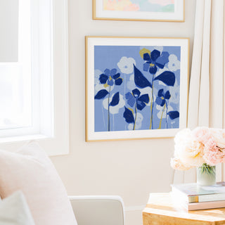 a cozy living room with a framed artwork of blue flowers hanging on the wall, a vase of pink flowers on a table, and a bookshelf filled with books.