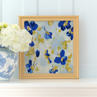 a framed painting of blue and yellow flowers on a white shelf, with a vase of white hydrangeas placed next to it.