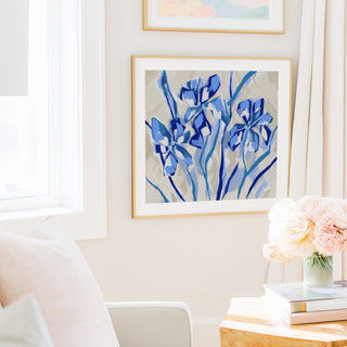 a cozy living room with a large abstract painting of blue flowers hanging on the wall, a white chair, a table with a vase of pink flowers, and a stack of books.