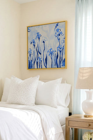 a serene bedroom scene with a bed adorned with white linens and a framed painting of blue flowers hanging above it. The painting adds a touch of color and life to the otherwise neutral-toned room.